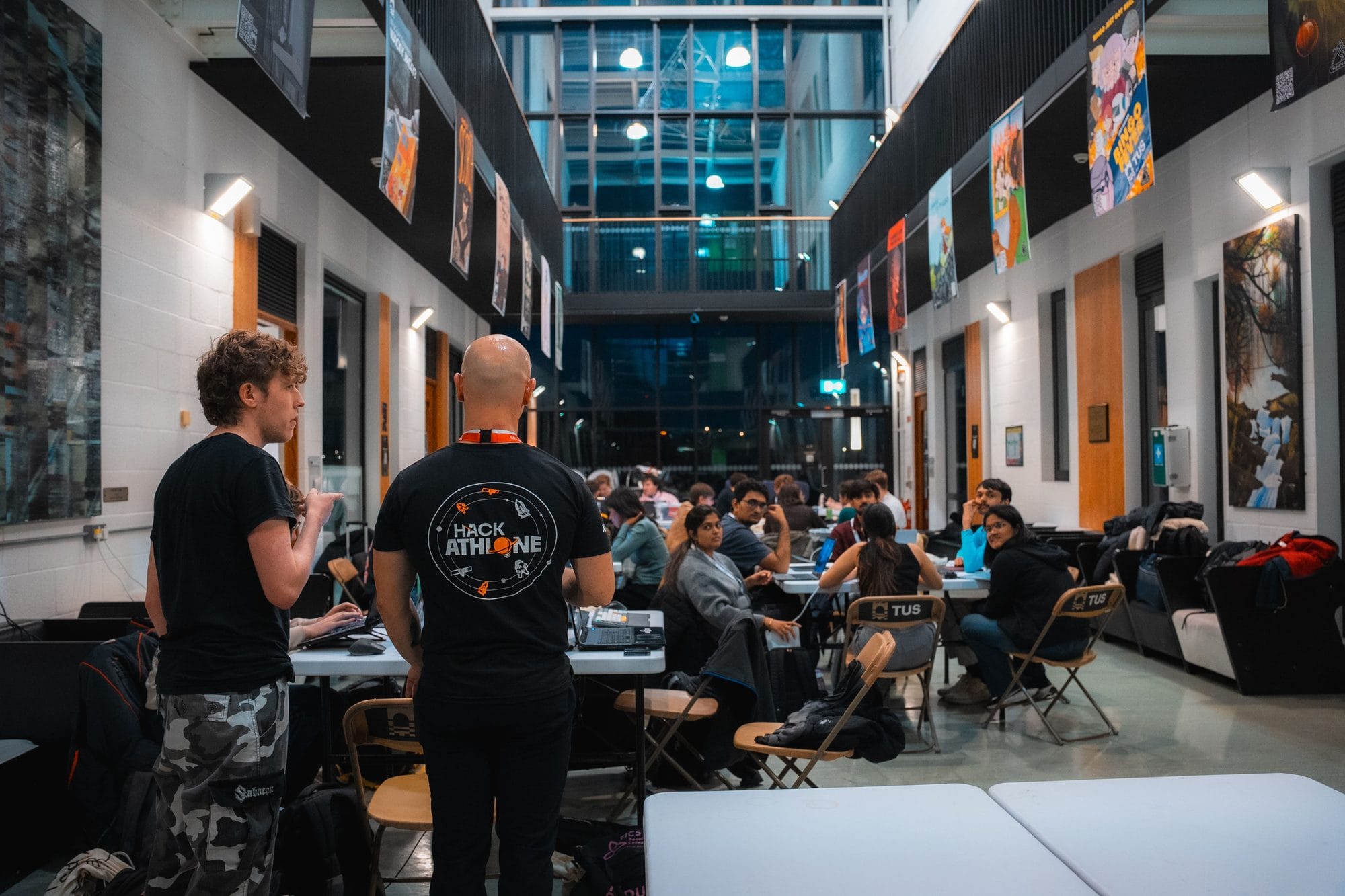 Late in the evening at HackAthlone 2025, two crew members look over the atrium as teams work at their tables