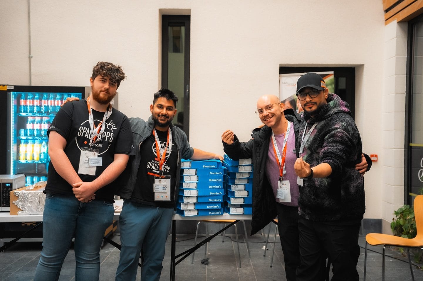 Four crew members with orange lanyards next to two stacks of pizza boxes at HackAthlone 2025